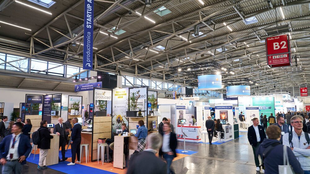 Blick in eine Messehalle während der productronica mit zahlreichen bunten Messeständen und vielen Besuchern, die sich dazwischen bewegen.