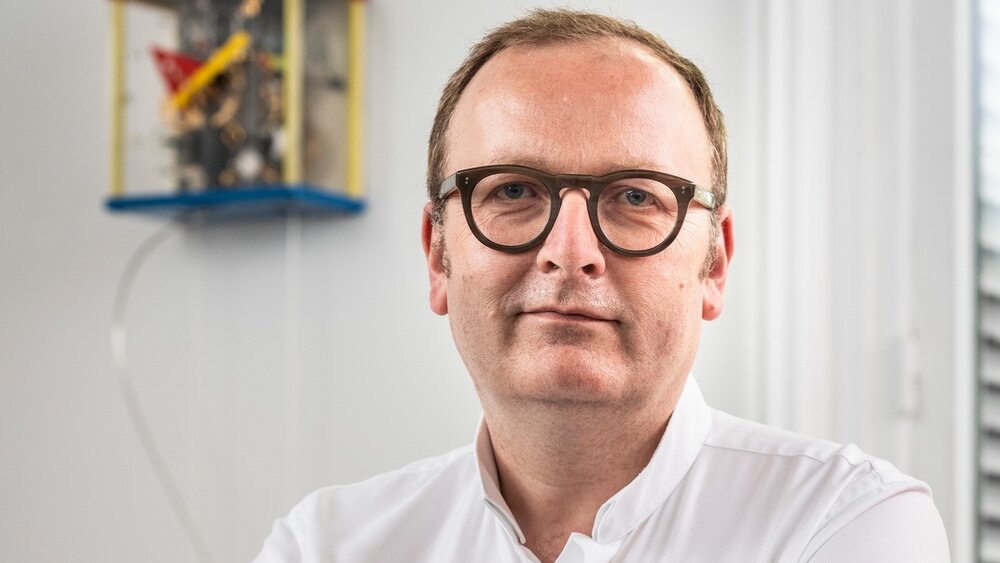Ein lächelnder Mann mit kurzen braunen Haaren trägt eine Brille mit breitem Rand, ein weißes Oberhemd und eine Armbanduhr.
