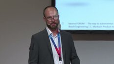 Ein Mann im Anzug spricht über ein Headset-Mikrofon am Konferenzpult vor einem Bildschirm.