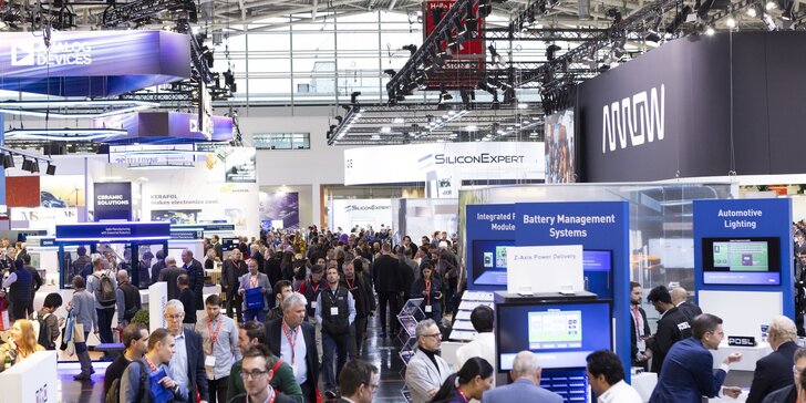 Large exhibition hall with numerous visitors walking between various booths and digital presentations at a trade fair.