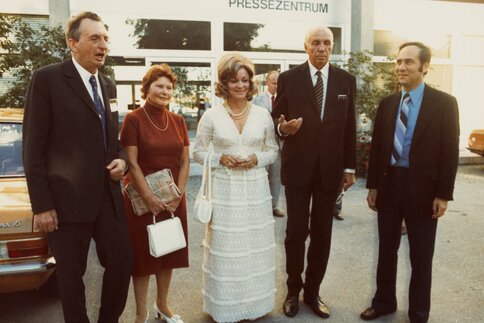 A group of five elegantly dressed people stand in front of a building labeled 