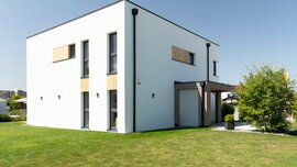 Modernes, weißes, zweistöckiges Haus mit großen Fenstern und Flachdach, umgeben von einem grünen Rasen.