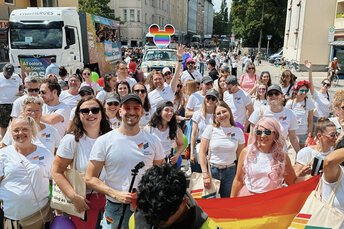 Eine Gruppe von Menschen bei einer Pride-Parade, die weiße Hemden mit Regenbogenfahnen tragen und eine große Regenbogenfahne halten.
