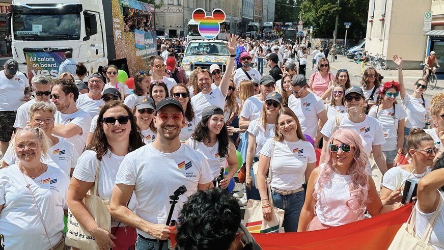 Eine Gruppe von Menschen bei einer Pride-Parade, die weiße Hemden mit Regenbogenfahnen tragen und eine große Regenbogenfahne halten.
