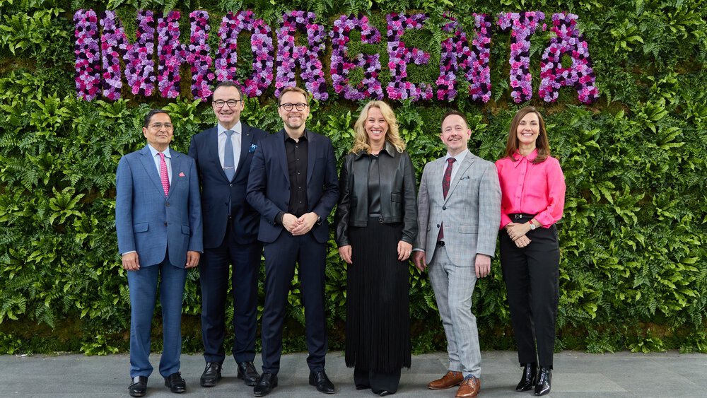 Six people in evening dress stand in front of a wall of grass with the floral inscription ‘INHORGENTA’.