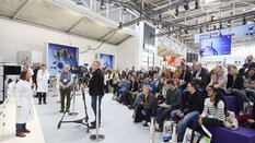 Publikum und ein Mann mit Kamera sehen einer Frau dabei zu, wie sie einen Vortrag hält