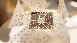 Ein Konstrukt aus dünnem, mit goldenen Schaltkreisen bedecktem Plastik. Helle dreieckige Formen gehen wie Blütenblätter von einem dunklen Quadrat in der Mitte aus.
