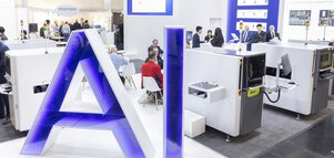 “AI” is written in large letters in blue and white on a platform in front of a trade fair stand at productronica. Behind it, groups of people are engrossed in conversation.