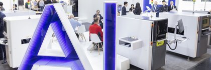 “AI” is written in large letters in blue and white on a platform in front of a trade fair stand at productronica. Behind it, groups of people are engrossed in conversation.