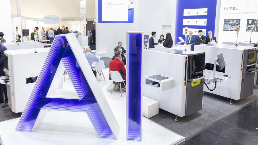 Große „AI“-Buchstaben in blau und weiß stehen vor einem Ausstellungsbereich auf der productronica. Hinter den Maschinen unterhalten sich mehrere Menschengruppen.