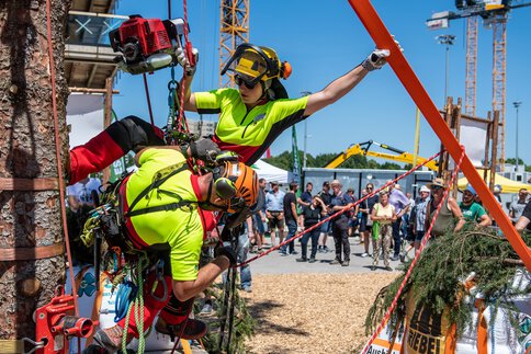  Zwei Personen in Schutzkleidung hängen gemeinsam neben einem Baumstamm und demonstrieren mit schwerem Gerät ihre Seilklettertechnik während der INTERFORST.