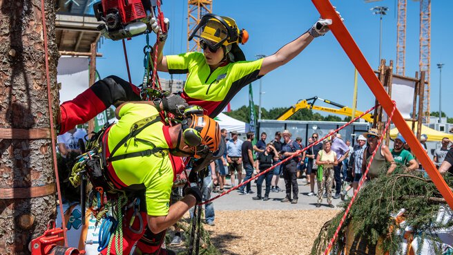Two people in protective clothing hang together next to a tree trunk and demonstrate their rope climbing technique with heavy equipment during INTERFORST.