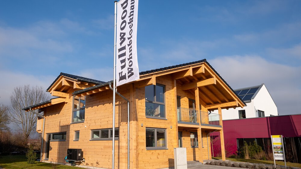Außenansicht eines zweistöckiges Holzhaus mit schrägem Dach, daneben ein Fahnenmast mit einem Banner des Herstellers.