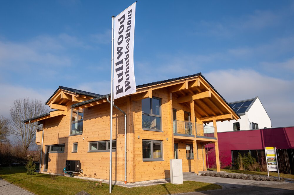 Außenansicht eines zweistöckiges Holzhaus mit schrägem Dach, daneben ein Fahnenmast mit einem Banner des Herstellers.