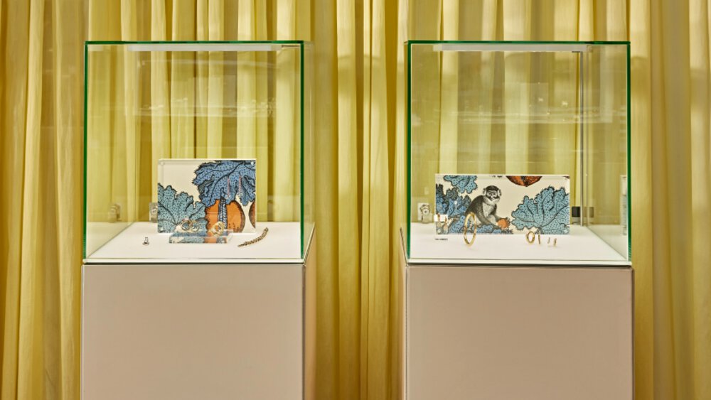 Two equally sized display cases containing gold jewelry and colorful nature pictures stand in front of a yellow curtain.