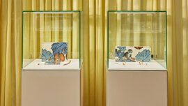 Two equally sized display cases containing gold jewelry and colorful nature pictures stand in front of a yellow curtain.