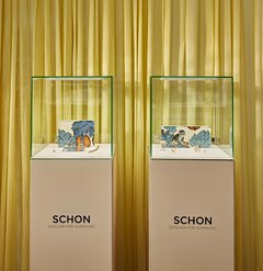 Two equally sized display cases containing gold jewelry and colorful nature pictures stand in front of a yellow curtain.