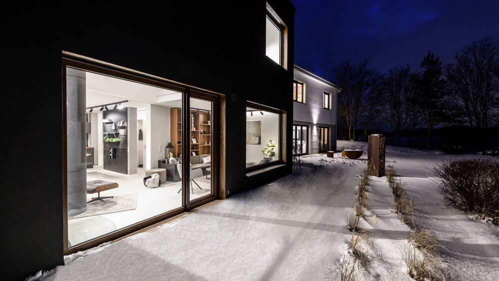Modernes Haus mit großen Fenstern, beleuchtetem Innenraum und schneebedecktem Weg bei Nacht.
