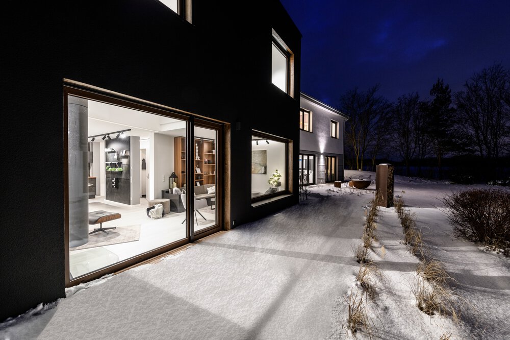 Modernes Haus mit großen Fenstern, beleuchtetem Innenraum und schneebedecktem Weg bei Nacht.
