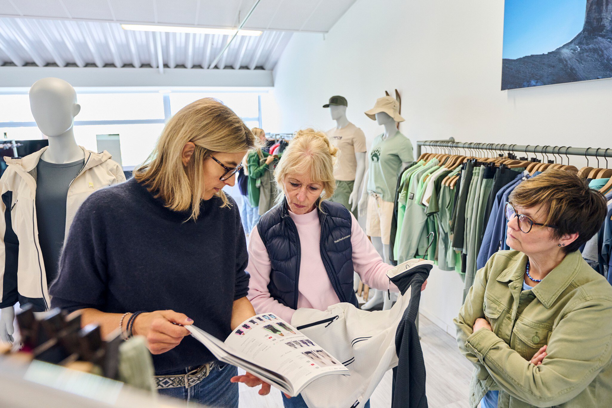 Drei Frauen stehen in einem Ausstellungsraum im MOC Ordercenter und betrachten ein weiß-graues Sportoberteil und einen Modekatalog.