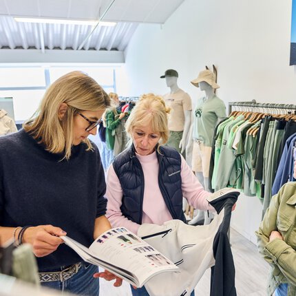 Drei Frauen stehen in einem Ausstellungsraum im MOC Ordercenter und betrachten ein weiß-graues Sportoberteil und einen Modekatalog.