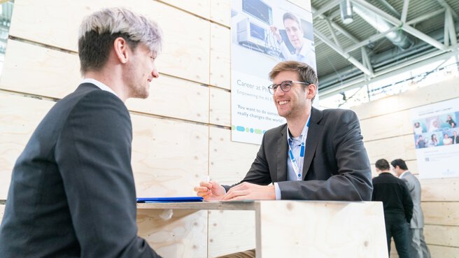 Zwei Personen in Geschäftskleidung unterhalten sich an einem Tisch, an einem Messestand mit Holzwänden.