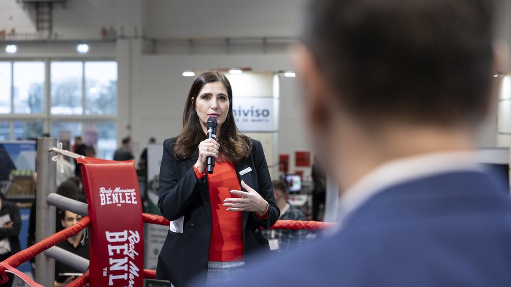 Eine Frau im Jackett und einem Mikro in der Hand steht in der Ecke eines Boxrings vor Publikum und spricht zu einem Mann im Ring.