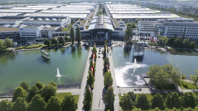 Luftaufnahme eines modernen Ausstellungszentrums mit Springbrunnen, Teichen, Bäumen und großen Gebäuden im Hintergrund.