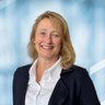Lächelnde Frau mit blondem Haar in einem dunklen Blazer und weißem Hemd vor einem blauen geometrischen Hintergrund.