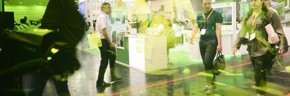 Messehalle mit laufenden Menschen, fotografiert durch eine farbige Scheibe, in der sich Ausstellungsstücke spiegeln.