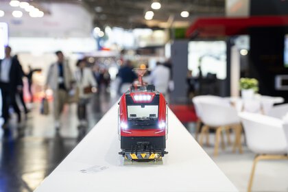 At transport logistic, a miniature model of a red railway stands on an elongated counter with blurred people in the background.