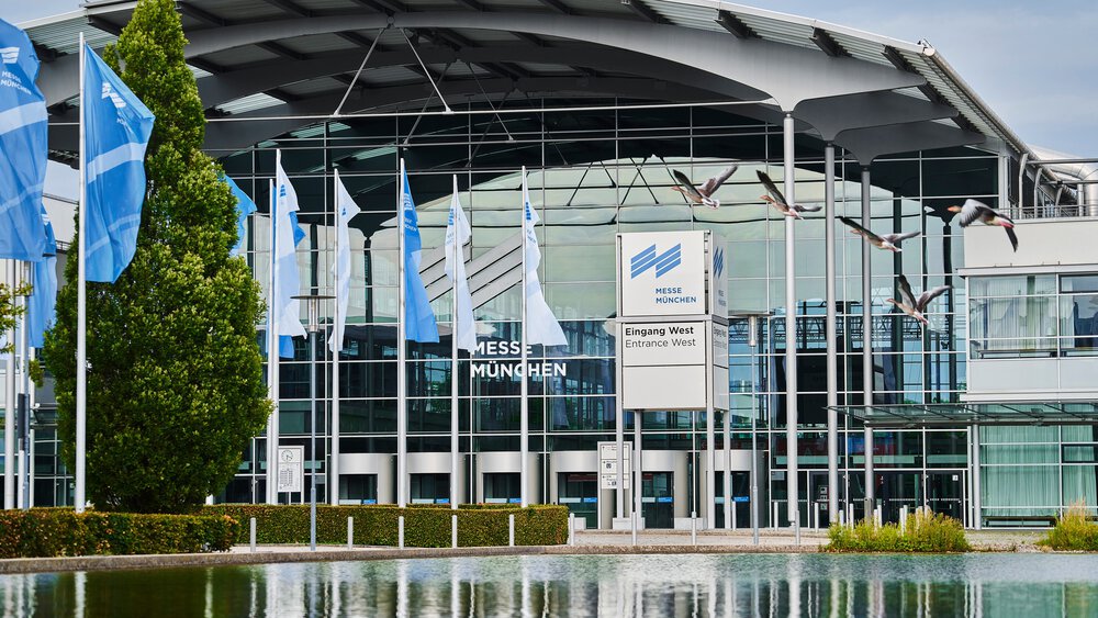 Moderner Eingang der Messe München mit blauen Fahnen, Glasfassade und fliegenden Vögeln über einem spiegelnden Becken.