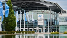 Moderner Eingang der Messe München mit blauen Fahnen, Glasfassade und fliegenden Vögeln über einem spiegelnden Becken.