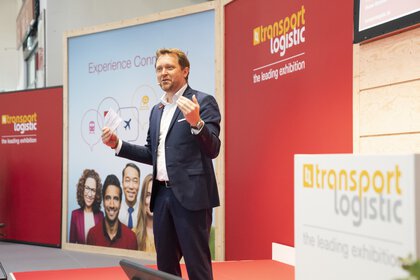 Ein Mann steht auf einer Bühne der transport logistic und spricht zu einem Publikum außerhalb des Bildes.