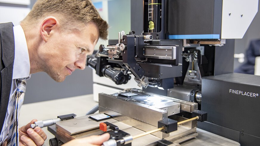 A man in a suit tests a complex, solid-looking production device with adjusting screws, magnification and lighting.