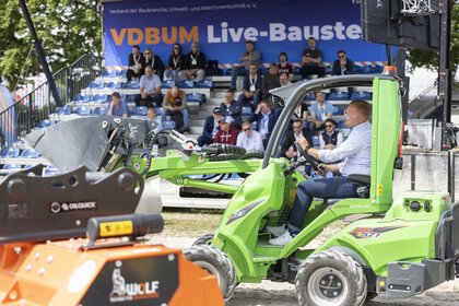 A trade fair visitor drives a small green excavator in the outdoor area in front of a grandstand with an audience.