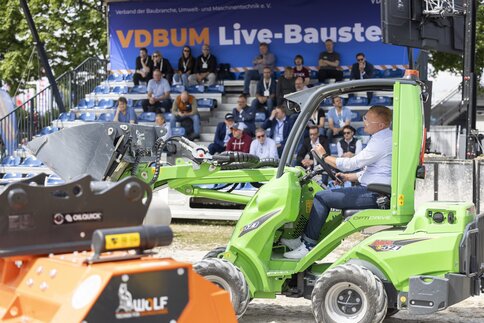 A trade fair visitor drives a small green excavator in the outdoor area in front of a grandstand with an audience.