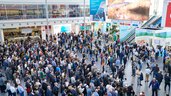 Erhöhte Ansicht einer sehr belebten, modernen Messehalle mit vielen Menschen, Ausstellungsständen und großen Bildschirmen.