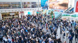 Elevated view of a very busy, modern exhibition hall with many people, exhibition stands, and large screens.