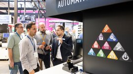 Vier Männer stehen gemeinsam an einem Stand mit einer bunt bedruckter Wand auf der productronica Messe und betrachten einen Computer.