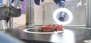 Ein Roboterarm mit einer Kamera und einem Ringlicht an der Spitze ist auf ein kleines oranges Modellauto auf einer dunklen Platte gerichtet.