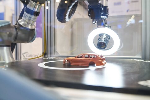 A robotic arm fitted with a camera and a ring light at the tip is pointed at a small orange model car on a dark surface.