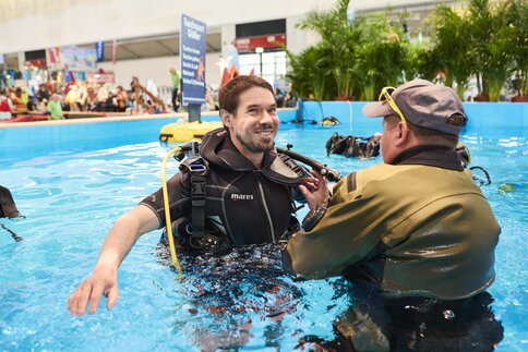 Ein männlicher Besucher der Mitmachfläche der free steht in Taucherausrüstung im Pool und bekommt die Ausrüstung erklärt und korrekt geprüft von einem Aussteller