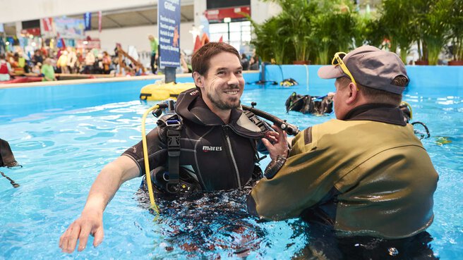 Ein männlicher Besucher der Mitmachfläche der free steht in Taucherausrüstung im Pool und bekommt die Ausrüstung erklärt und korrekt geprüft von einem Aussteller