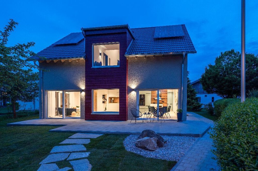 Modernes zweistöckiges Haus mit großen Fenstern, Terrassenmöbeln und einem Gartenweg in der Dämmerung.