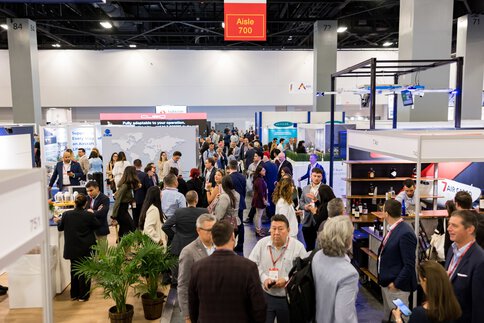 Numerous trade fair visitors stand in small groups at several exhibition stands in an event hall and engage in lively conversation.