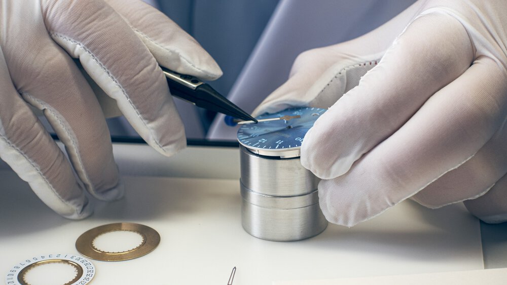Close-up of two hands in white gloves using tweezers to insert a hand into a blue dial, while other watch parts lie on the table.