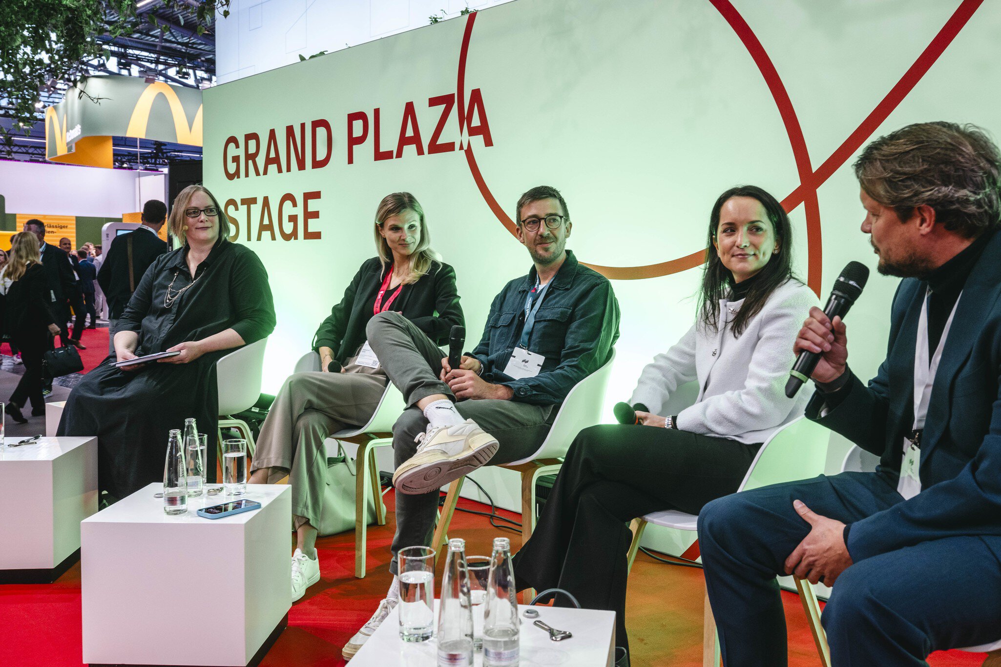 A panel discussion on the “Grand Plaza Stage” at EXPO REAL: Five people sit in a row on white chairs. The man on the right is speaking into a microphone, while the others listen and smile. Small tables with water bottles and glasses are placed in front of them. In the background, trade fair visitors walk through the exhibition hall.