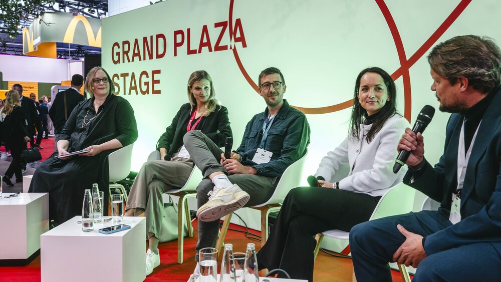 A panel discussion on the “Grand Plaza Stage” at EXPO REAL: Five people sit in a row on white chairs. The man on the right is speaking into a microphone, while the others listen and smile. Small tables with water bottles and glasses are placed in front of them. In the background, trade fair visitors walk through the exhibition hall.
