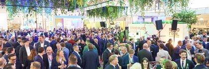 gefüllte Messe- oder Eventhalle mit vielen Menschen in Businesskleidung, im Vordergrund Stehtische, darüber grüne Pflanzeninstallation.